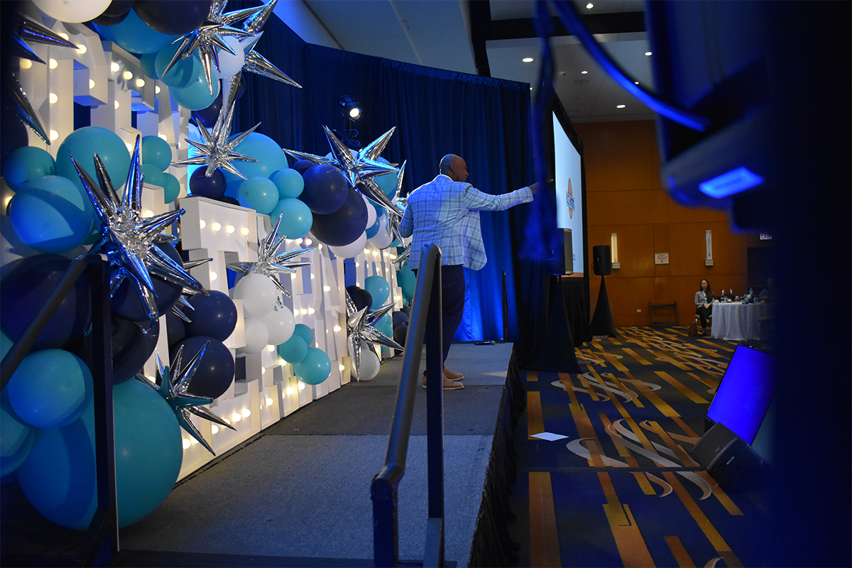 A man stands on stage giving a presentation, surrounded by blue and white balloons and silver star decorations, in a large conference room.