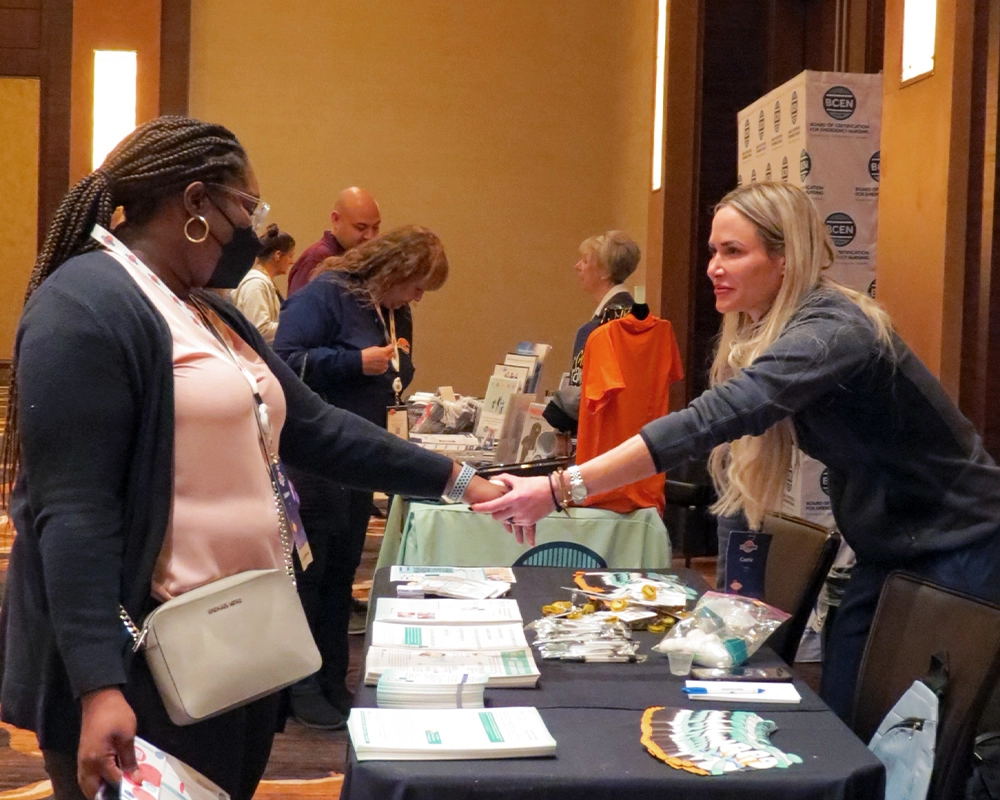 Two women shake hands across a table at a conference or expo, with informational materials and promotional items displayed in front of them. Other attendees interact at nearby tables.
