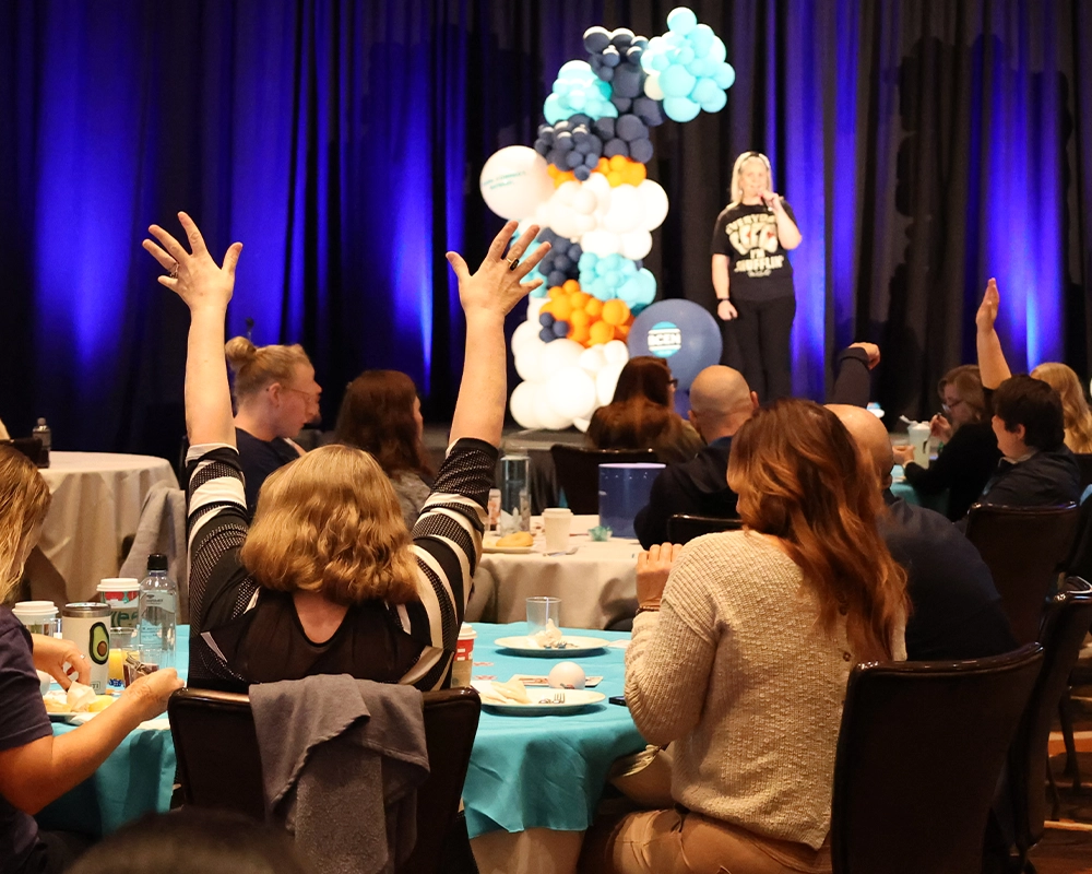 A woman raises her arms while seated at a round table during an indoor event with a speaker on stage and balloon decorations in the background.