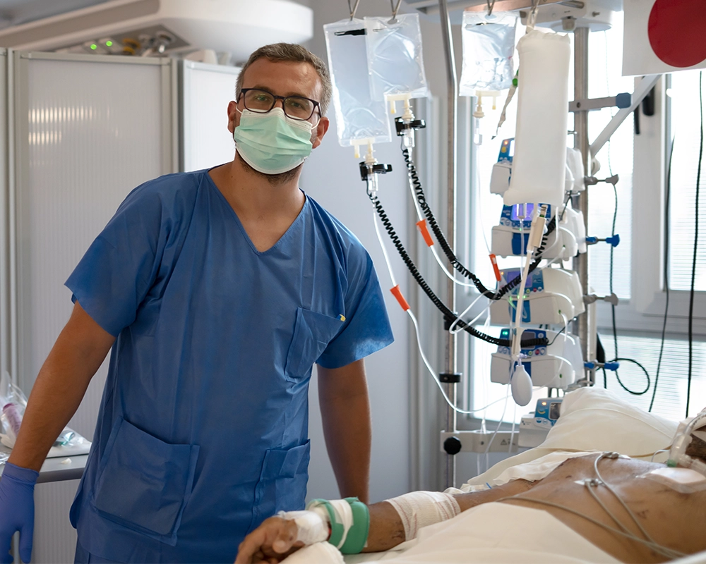 A healthcare worker in blue scrubs and a mask stands next to a patient connected to medical equipment in a hospital room.