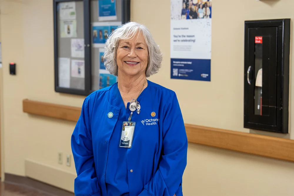 A woman in a blue Ochsner Health RN uniform stands and smiles in a hospital hallway, with medical posters and equipment visible on the wall behind her.