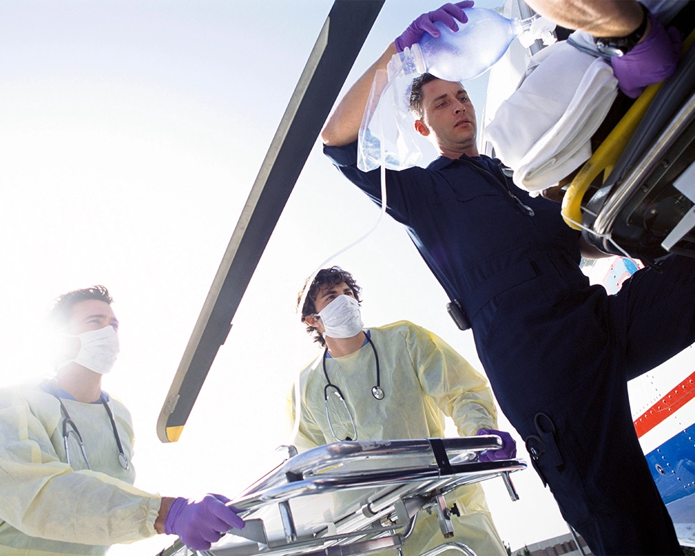 Three medical professionals wearing protective gear move a patient on a stretcher toward a helicopter, with one holding an IV bag.
