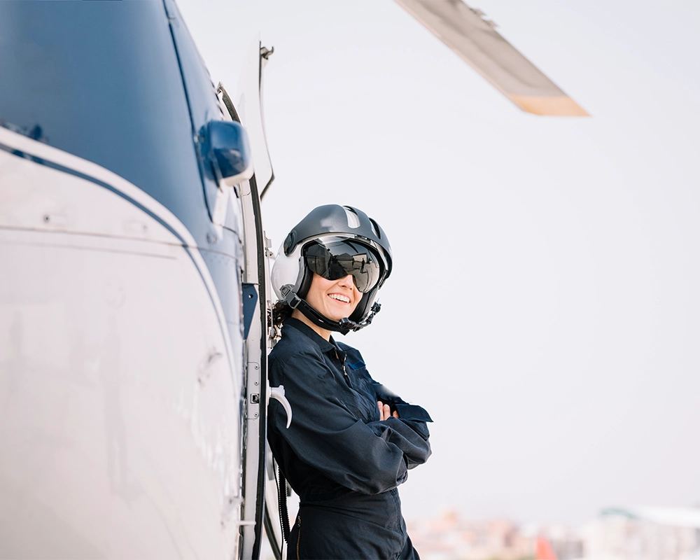 A person wearing a helmet and flight suit stands with arms crossed, leaning against a helicopter on a clear day.