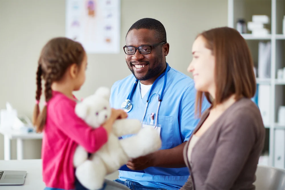 A doctor in scrubs smiles while talking to a young girl holding a teddy bear and an adult woman, likely her mother, in a medical office.