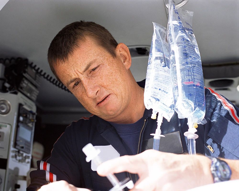 A paramedic prepares an IV line next to two hanging IV fluid bags inside an emergency vehicle.