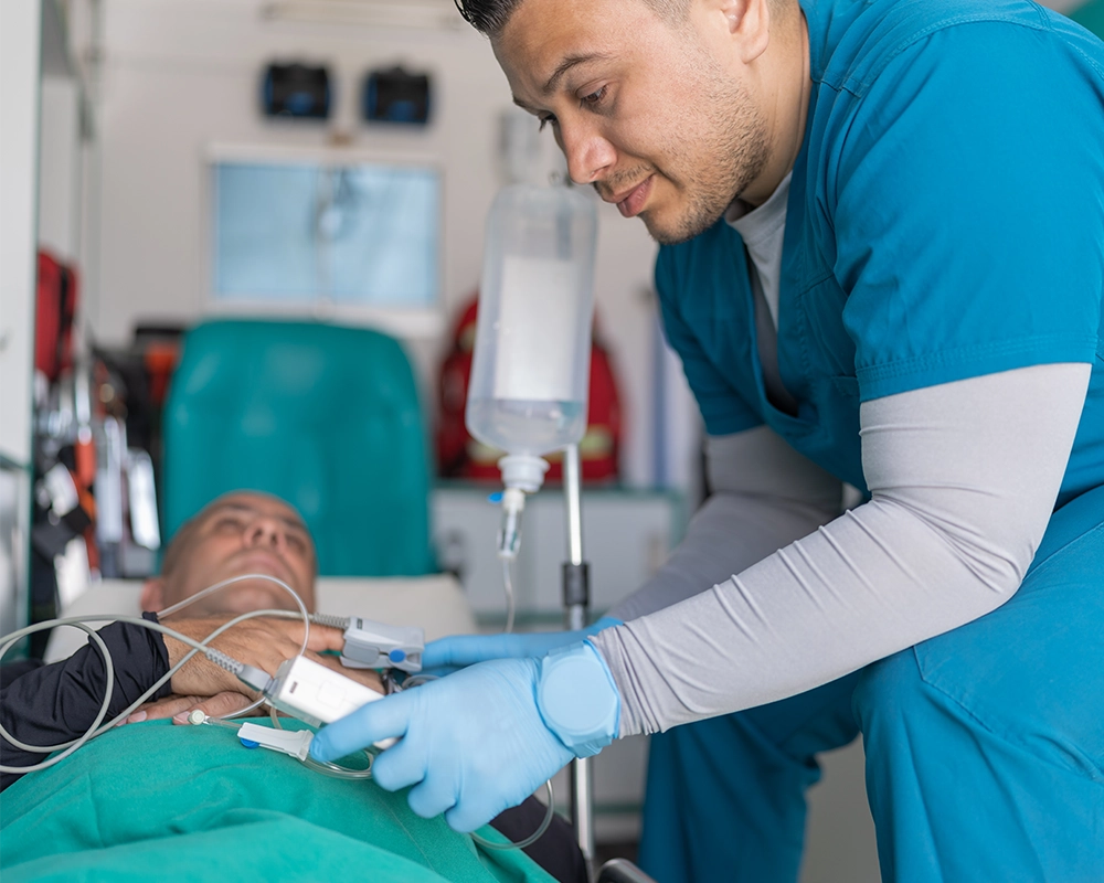 A healthcare worker in blue scrubs and gloves monitors a patient lying on a stretcher with medical equipment in a clinical setting.