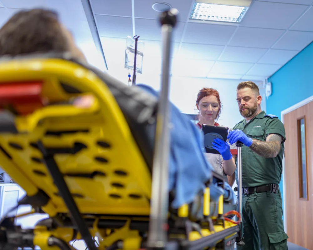 A patient lies on a stretcher while two medical professionals, one holding a tablet, stand nearby in a brightly lit hospital room.