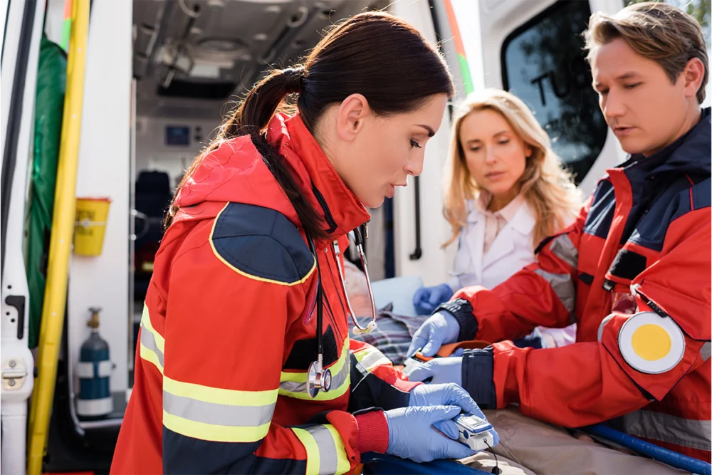 Three paramedics in red uniforms provide medical assistance to a patient inside an ambulance, using monitoring equipment and medical supplies.
