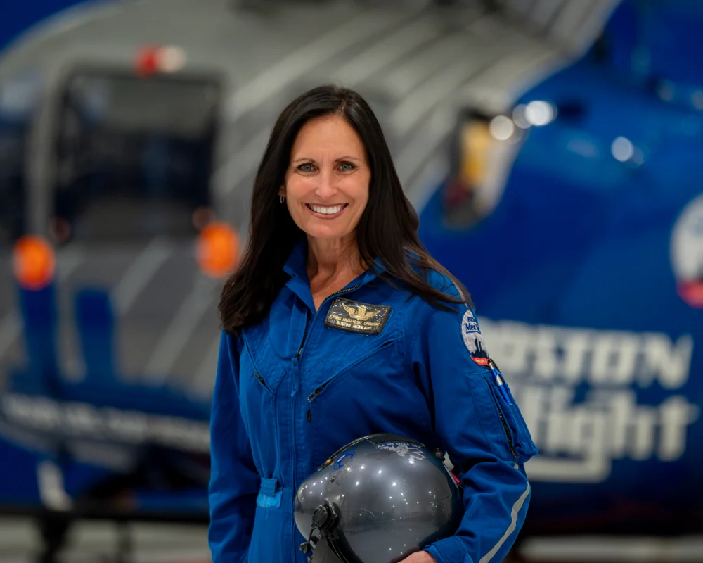 A woman in a blue flight suit, 2026 Distinguished CFRN Award Winner, holds a helmet and stands in front of a blue helicopter with “Boston MedFlight” visible on the side.