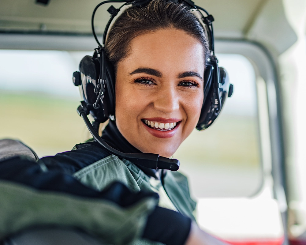 A woman wearing a headset and pilot uniform smiles while seated inside an aircraft cockpit.