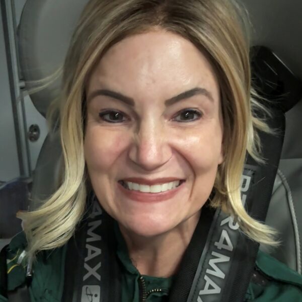 A woman with blonde hair smiles at the camera while seated and wearing a green uniform and a harness labeled "PARA MAX.