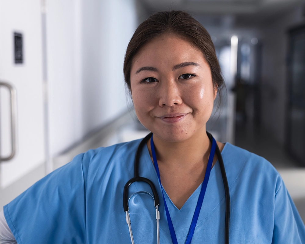 A healthcare worker in blue scrubs and a stethoscope stands in a hospital corridor, looking at the camera with a slight smile.