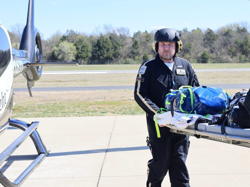 Two paramedics in flight suits and helmets transport a patient on a stretcher to a medical helicopter on a sunny day.