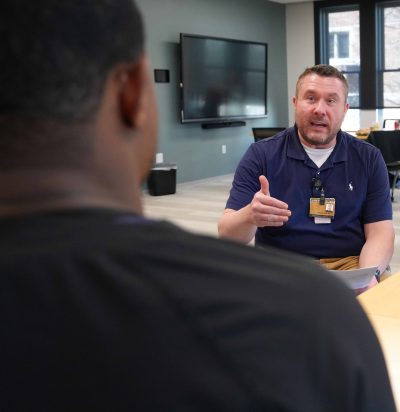 A man in a navy polo shirt speaks to another person across a table in a modern office or meeting room.