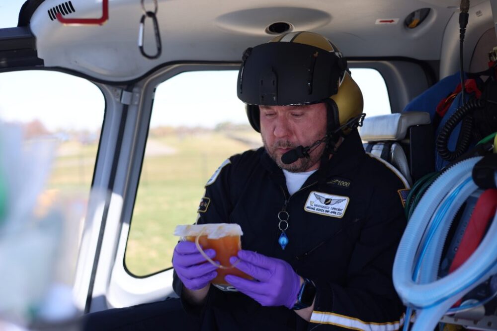 A flight paramedic wearing a helmet and gloves examines a bag of blood inside a medical helicopter.