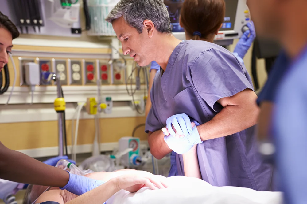 Medical professionals attend to a patient in a hospital setting, with one doctor holding the patient’s arm and others providing care, surrounded by medical equipment.