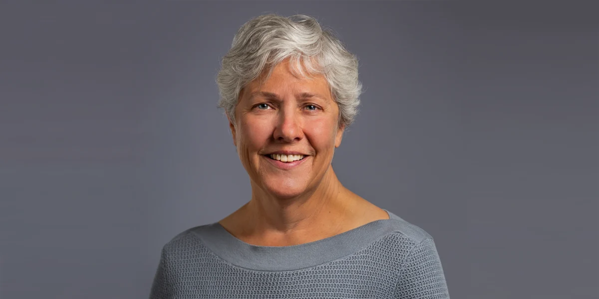 Older woman with short gray hair, wearing a light gray top, smiling in front of a plain gray background.