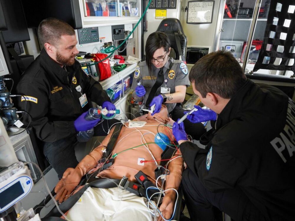 Three paramedics practice emergency medical procedures on a training mannequin inside an ambulance, surrounded by medical equipment.