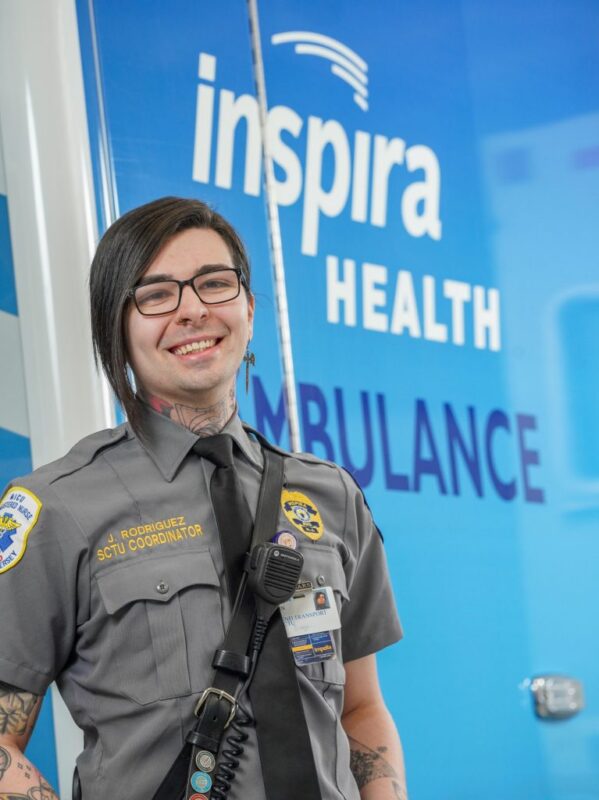A person in a gray EMT uniform, wearing glasses and a radio, stands smiling in front of an Inspira Health ambulance sign.