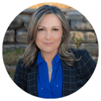 Woman with shoulder-length light brown hair wearing a blue blouse and black checkered blazer, sitting outdoors with a stone wall in the background.