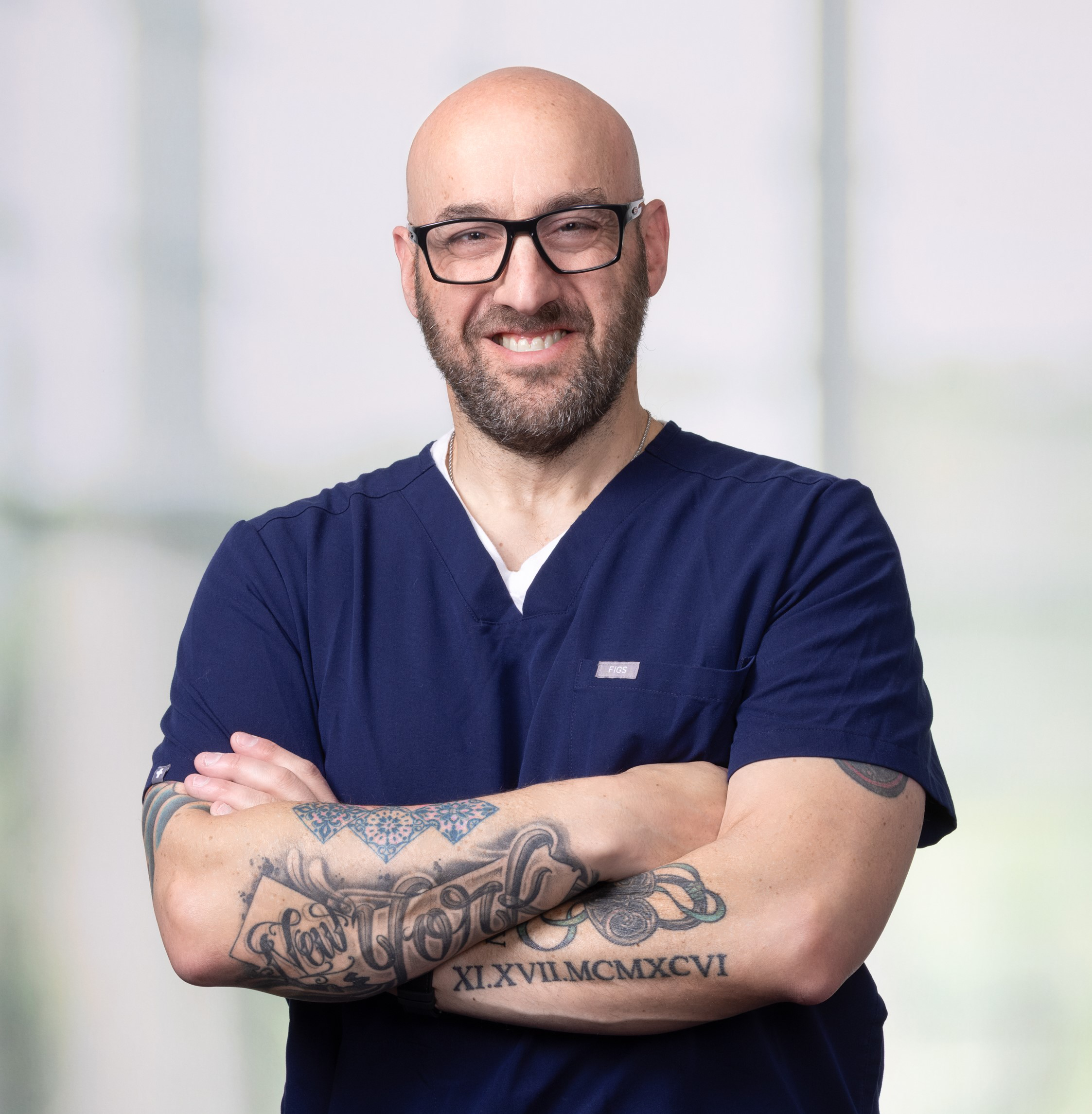 Bald man with glasses and tattoos on his arms, wearing navy blue scrubs, stands with arms crossed, smiling in front of a blurred light background—the proud 2026 Distinguished CEN Award Winner.