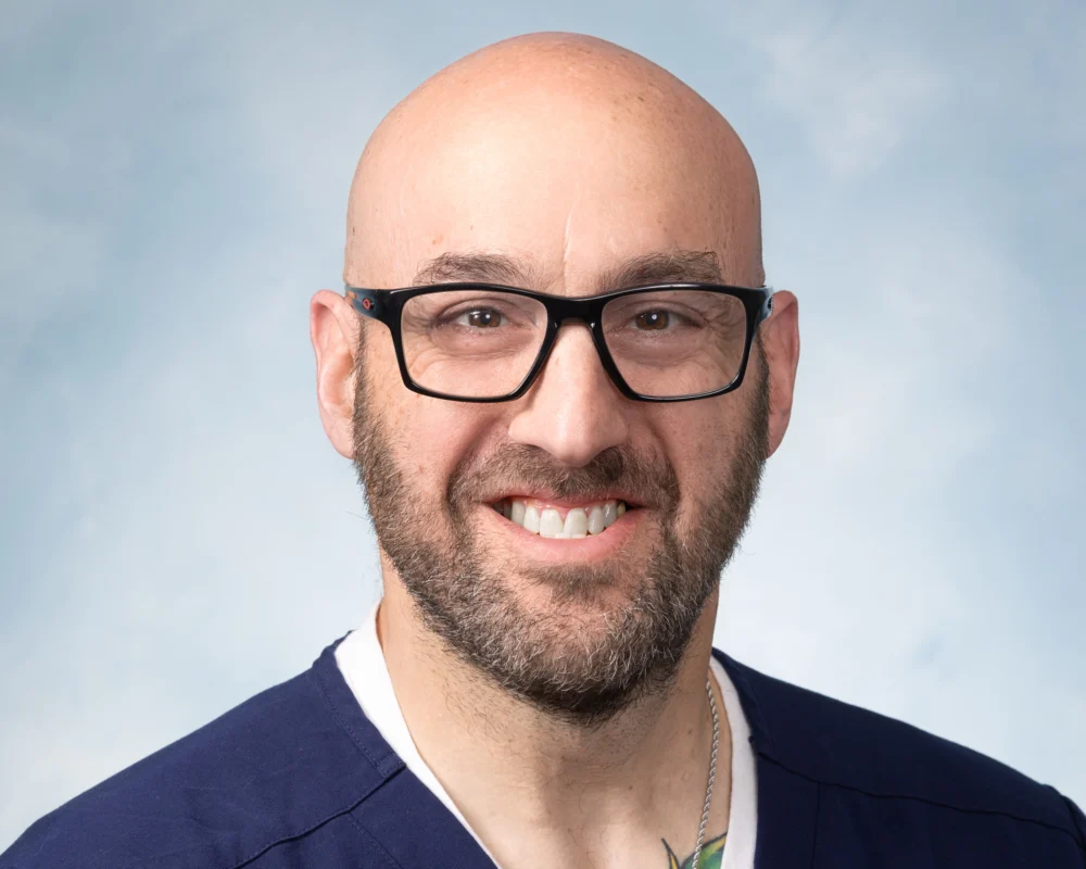 A bald man with a beard and glasses smiles at the camera, wearing a dark blue shirt and a white undershirt, against a light blue background. He is the 2026 Distinguished CEN Award Winner.