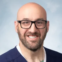 A bald man with a beard and glasses smiles at the camera, wearing a dark blue shirt and a white undershirt, against a light blue background. He is the 2026 Distinguished CEN Award Winner.