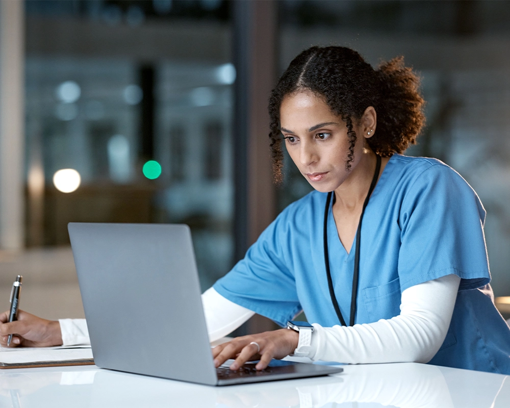 A person in medical scrubs sits at a desk, typing on a laptop and writing notes with a pen.