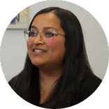 A woman with long dark hair and glasses smiles while seated indoors against a light-colored background, celebrating Certified Nurses Day.
