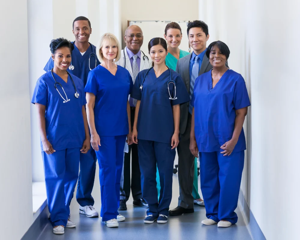 A diverse group of eight healthcare professionals, including doctors and nurses, stand together in a hospital hallway, facing the camera and smiling.