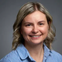 Woman with shoulder-length blonde hair wearing a blue and white striped shirt, smiling at the camera against a plain gray background. She is the proud recipient of the 2026 Distinguished CPEN Award.