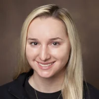 A woman with straight blonde hair, wearing a black zip-up top and a necklace, smiles in front of a plain brown background—2026 Distinguished CBRN Award Winner.