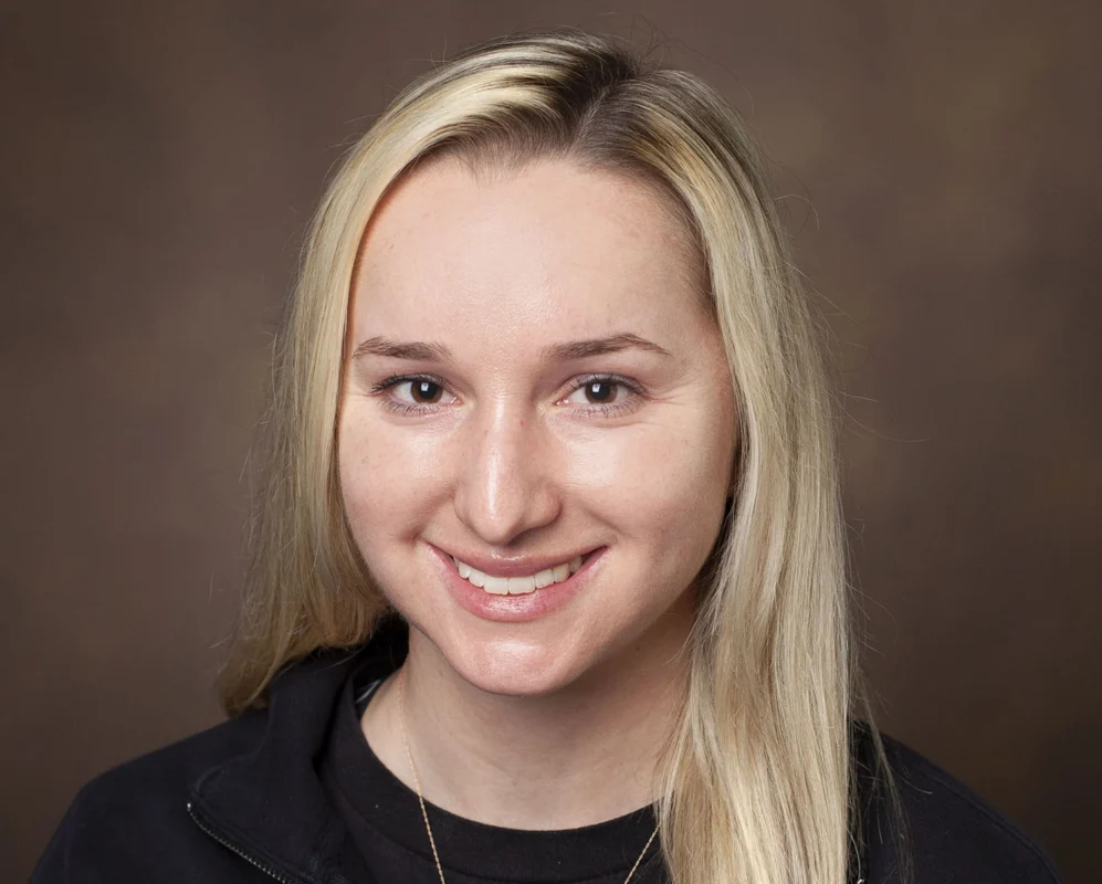 A woman with straight blonde hair, wearing a black zip-up top and a necklace, smiles in front of a plain brown background—2026 Distinguished CBRN Award Winner.