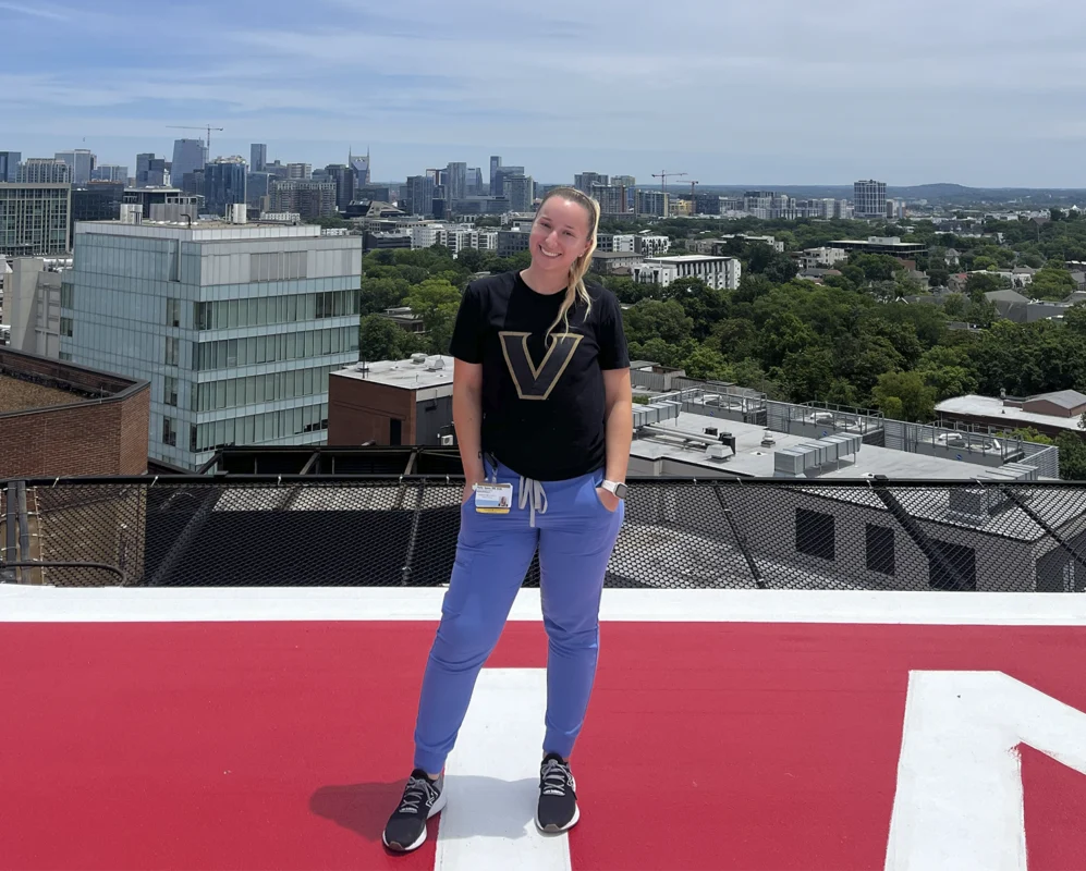 Person stands on a rooftop helipad with a city skyline and scattered clouds in the background, embodying the poise of a 2026 Distinguished CBRN Award Winner.
