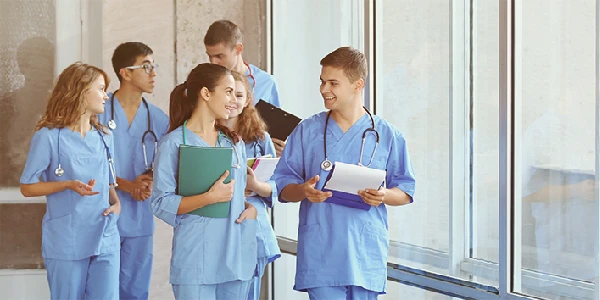 A group of medical students in scrubs walk down a hallway, holding folders and clipboards, and talking to each other.
