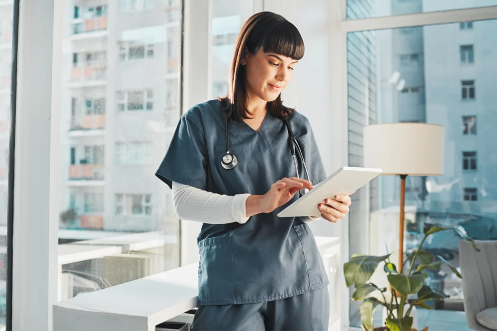 A healthcare professional in scrubs with a stethoscope uses a tablet in a bright, modern office with large windows.