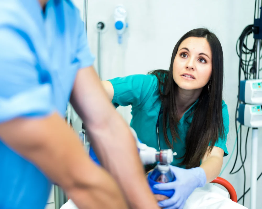 A healthcare worker in scrubs and gloves assists another person with a manual resuscitator in a medical setting.