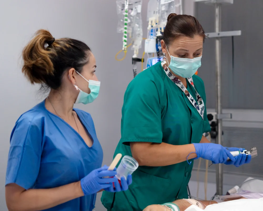 Two healthcare workers in scrubs and masks attend to a patient, preparing medical equipment and holding sample containers in a clinical setting.