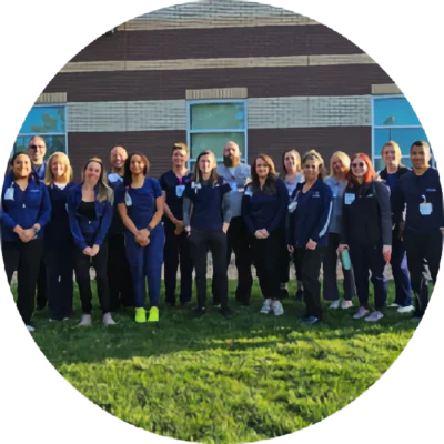 A group of sixteen people in blue uniforms stand in a row on grass in front of a brick building, facing the camera.