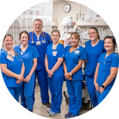 A group of seven healthcare professionals in blue scrubs pose together and smile in a hospital room with medical equipment in the background.