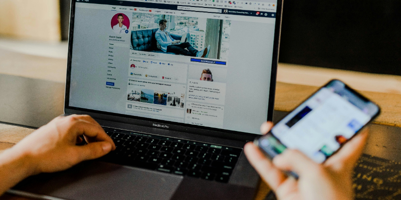 Person using a laptop to view a social media profile while holding a smartphone, both screens displaying online content.