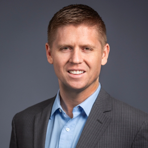A man with short light brown hair wearing a gray suit jacket and light blue shirt, smiling against a plain gray background.
