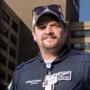 A man in a medical uniform with an ID badge stands outside in front of tall buildings, wearing sunglasses on his cap.