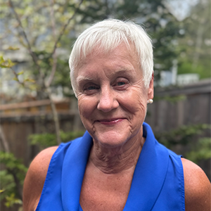 Older woman with short white hair and a blue sleeveless top smiles outdoors in a backyard setting.