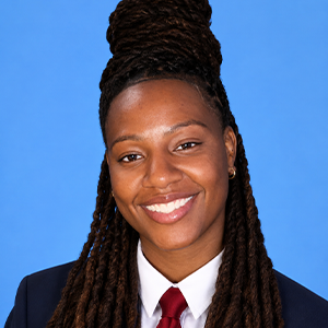 A person with long, twisted hair styled in a high bun, wearing a dark suit, white shirt, and red tie, smiles in front of a plain blue background.