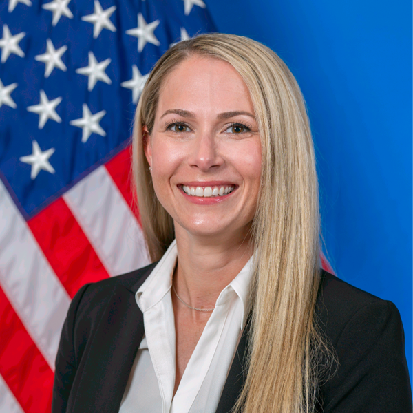 A woman with long blonde hair in a suit jacket and white shirt smiles in front of a U.S. flag and a blue background.