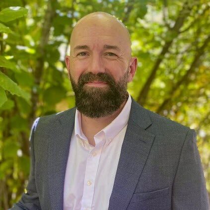 A man with a bald head and beard, wearing a gray suit jacket and white shirt, stands outdoors in front of green leafy trees.