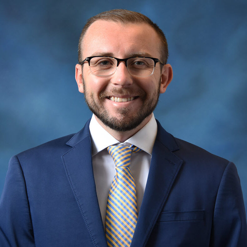 Man in a blue suit, light blue shirt, and yellow striped tie standing in front of a blue background, smiling at the camera.