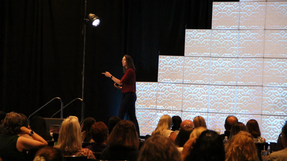 A woman stands on stage speaking to an audience, with geometric patterned panels stacked in a stepped design behind her.