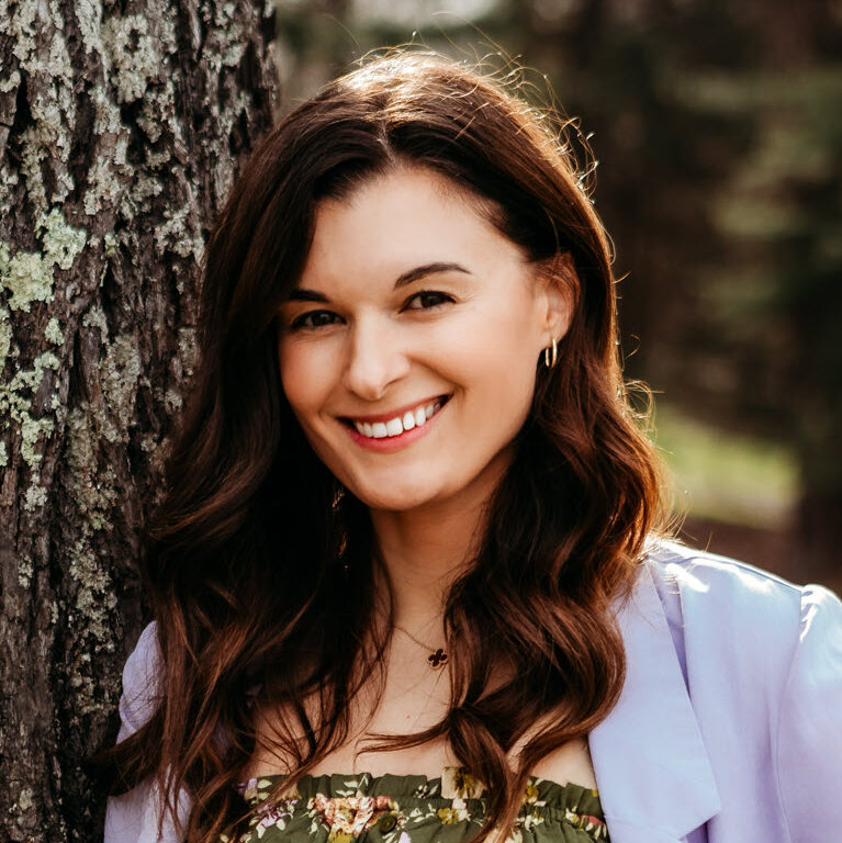 A woman with long brown hair wearing a floral dress and a light purple blazer stands outdoors, leaning against a tree and smiling at the camera.